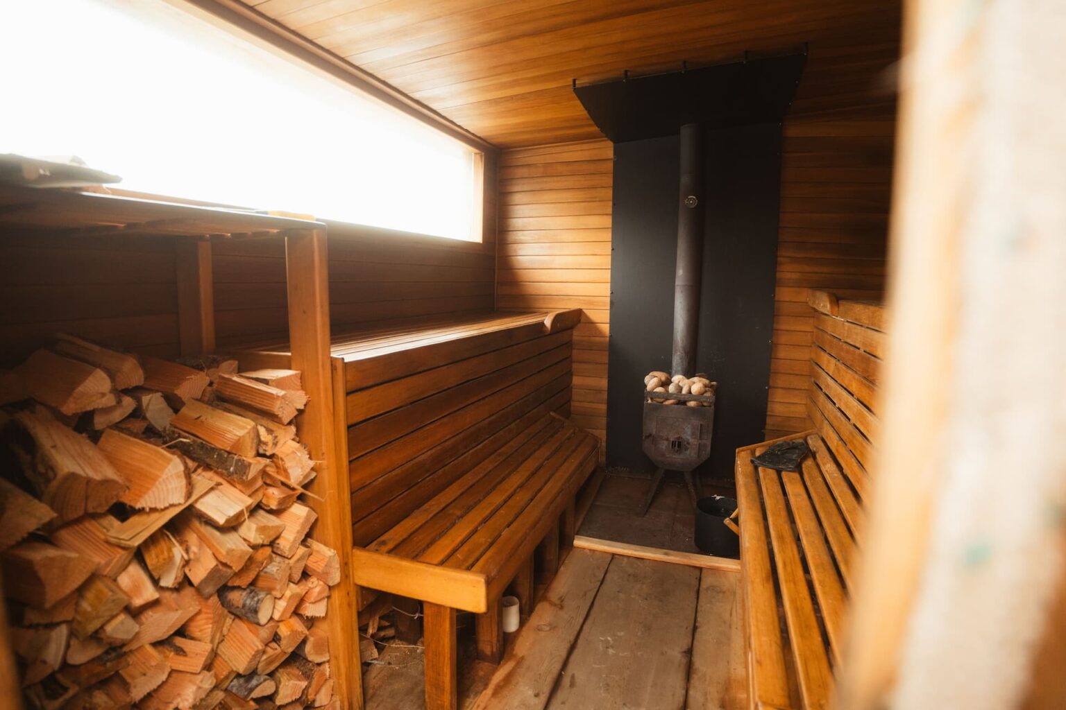 Puinen sauna, jossa lauteet, halkopino ja puukiuas kapean ikkunan alla