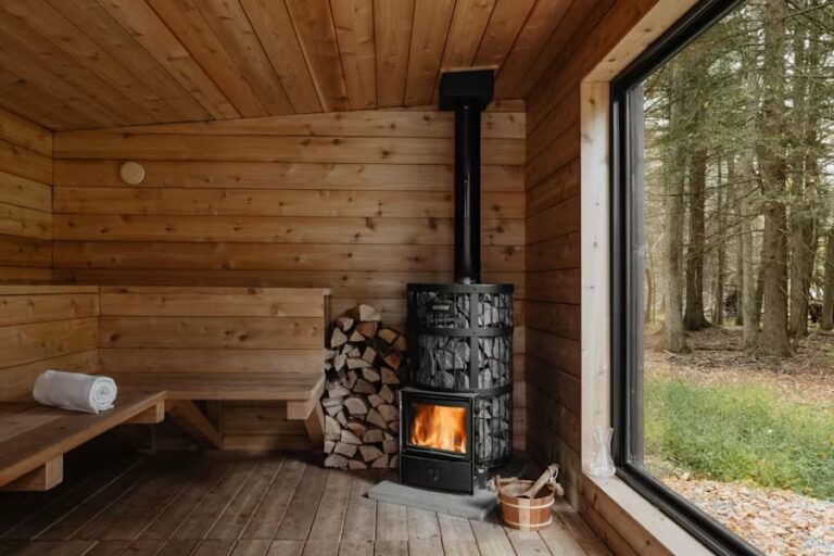 Cozy wooden sauna with a lit wood stove, stacked firewood, a bench, and a large window looking out to a forest.