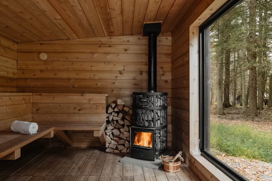 Cozy wooden sauna with a lit wood stove, stacked firewood, a bench, and a large window looking out to a forest.